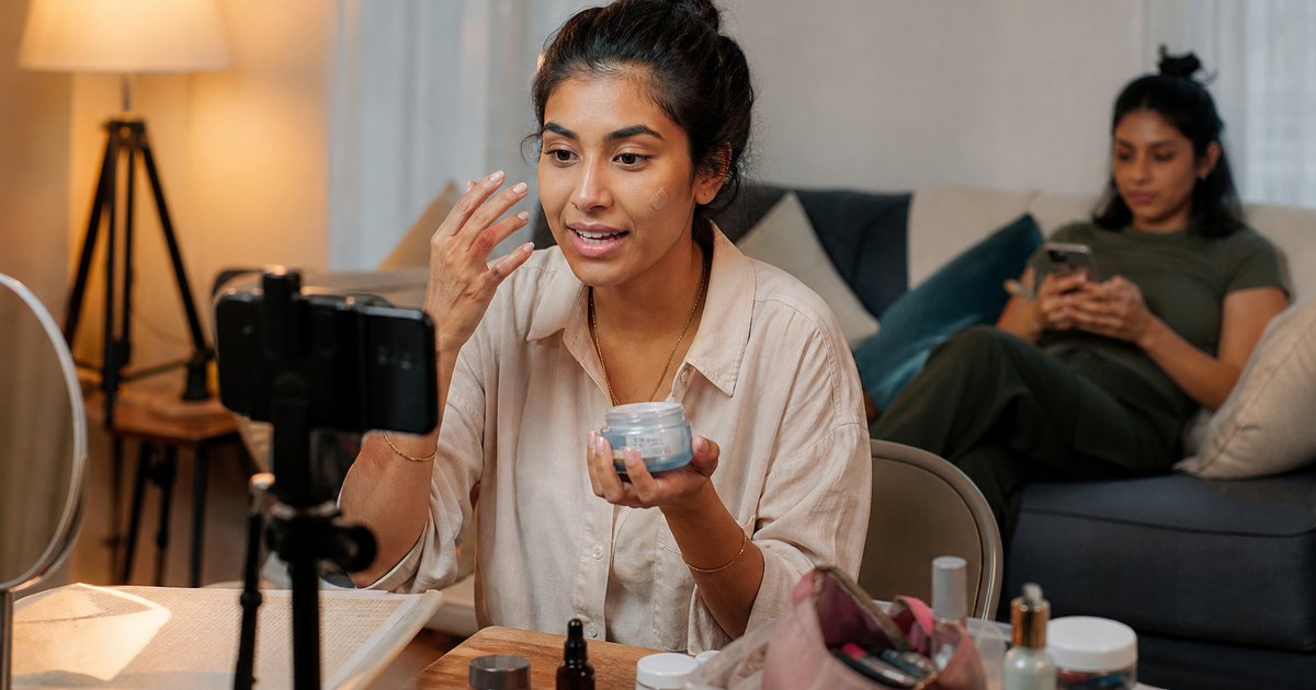 Creator filming a get-ready-with-me video in a shared apartment living room while a friend sits on the couch in the background scrolling a phone, warm lamp light