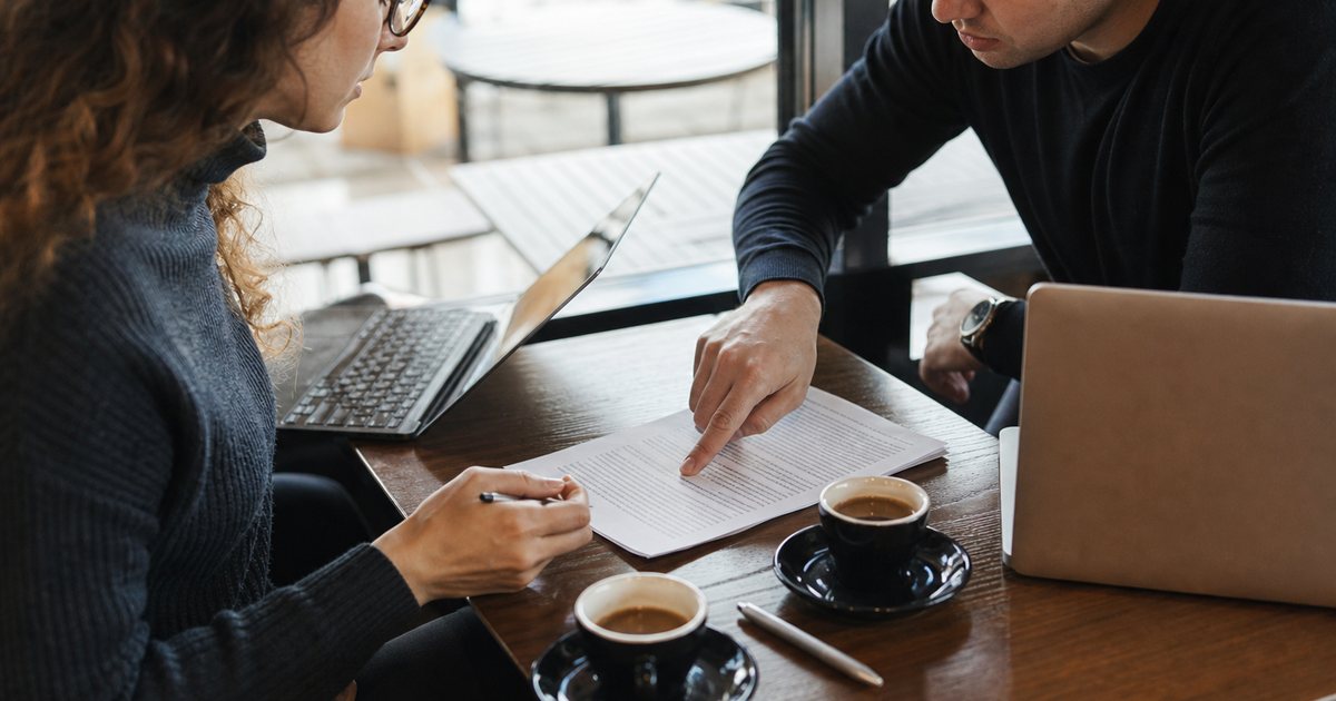 Two people at a cafe table with laptops and a printed one-page agreement between them, one pointing at a line on the page, espresso cups and a pen nearby